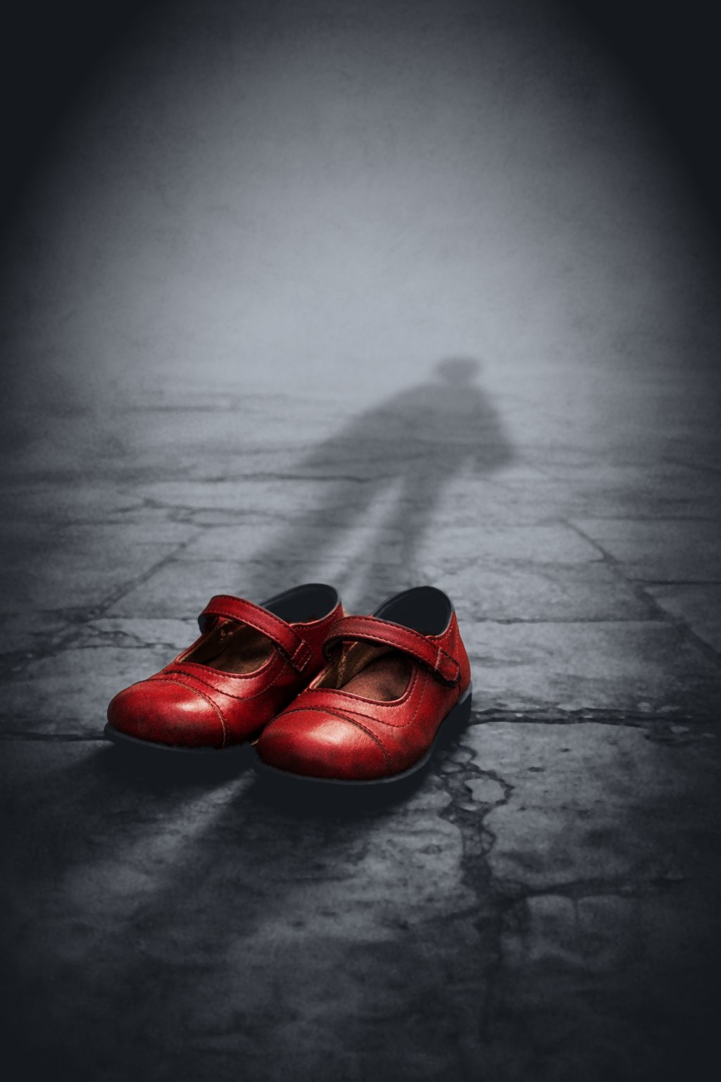A pair of worn red children’s shoes on grey stone, with the shadow of a child falling across the ground behind them